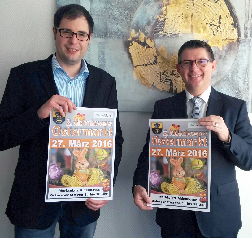 Bürgermeister Ralf Claßen (r.) und sein Mitarbeiter Marcel Dederichs präsentieren das offizielle Plakat zum 2. Aldenhovener Ostermarkt.