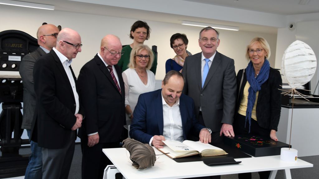 Foto „Johann Lafer Eintrag Goldenes Buch“: Johann Lafer, Träger des Dürener Papiermacherordens, trug sich im Papiermuseum in das Goldene Buch der Stadt ein, rechts auf dem Tisch der diesjährige elegante weiße Papiermacherorden mit Sockel. Foto: Stadt Düren