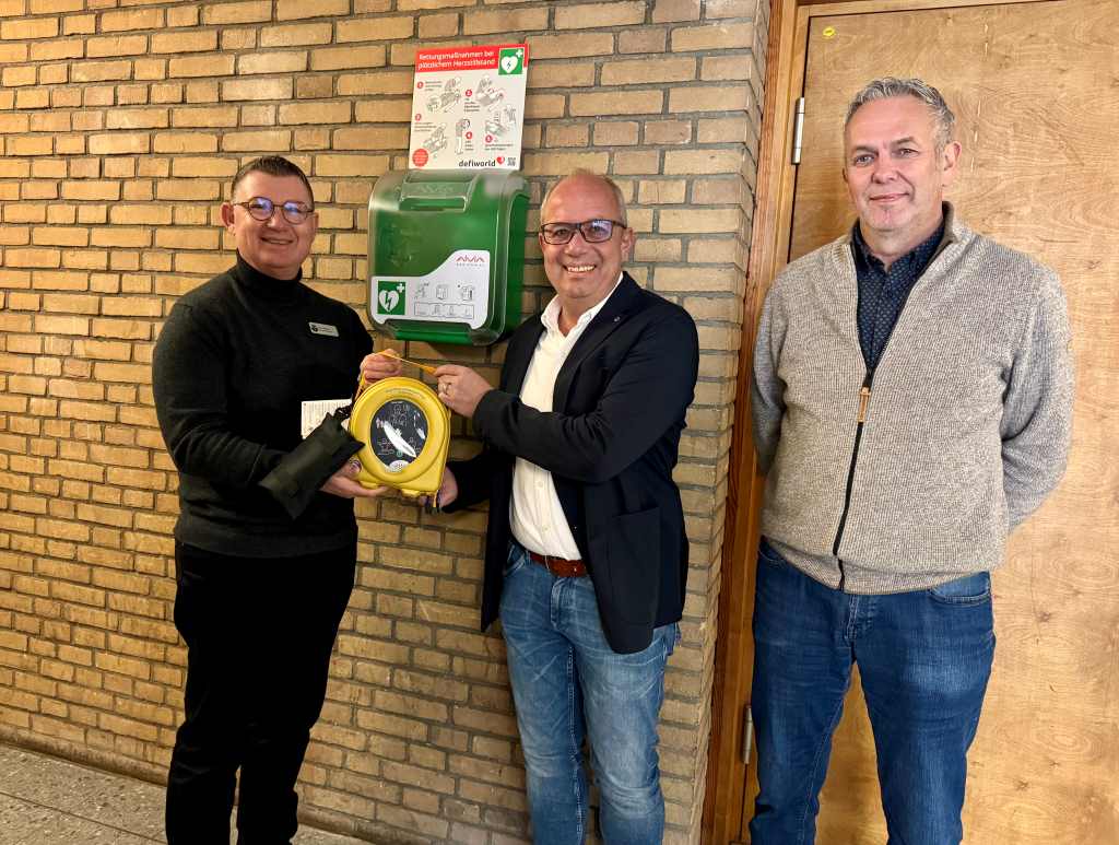 Freuen sich über den neuen AED für das Schul- und Vereinssportzentrum in der Aldenhovener Schwanenstraße aus der Hand von ENGIE-Niederlassungsleiter Sascha Strehl (2.v.l.): Gemeindebrandmeister Wilfried Thelen (rechts) und Bürgermeister Ralf Claßen (links) 